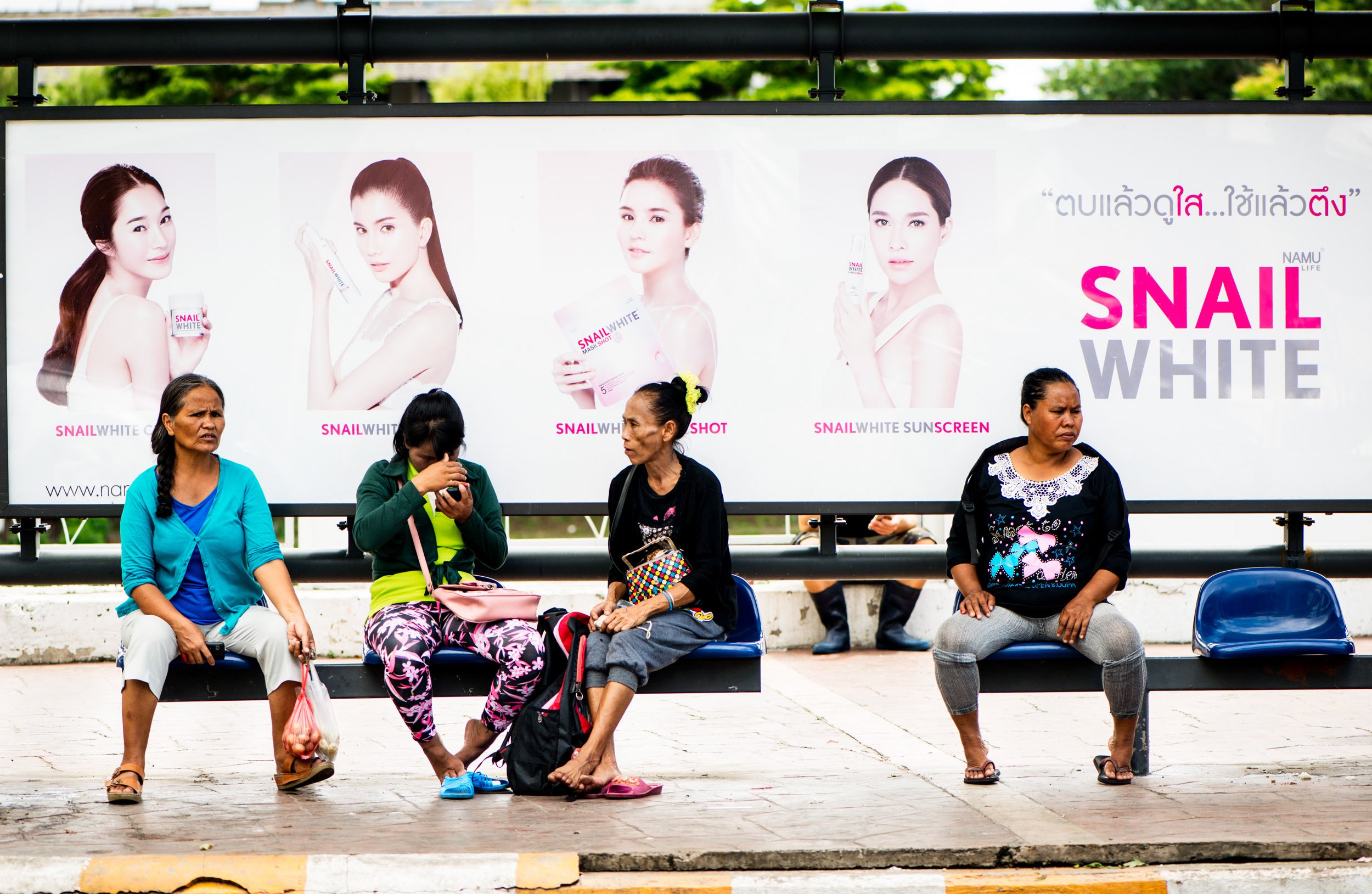 Women sitting at a bus stop with an advertisement for skin whitening © Kevin Landwer-Johan